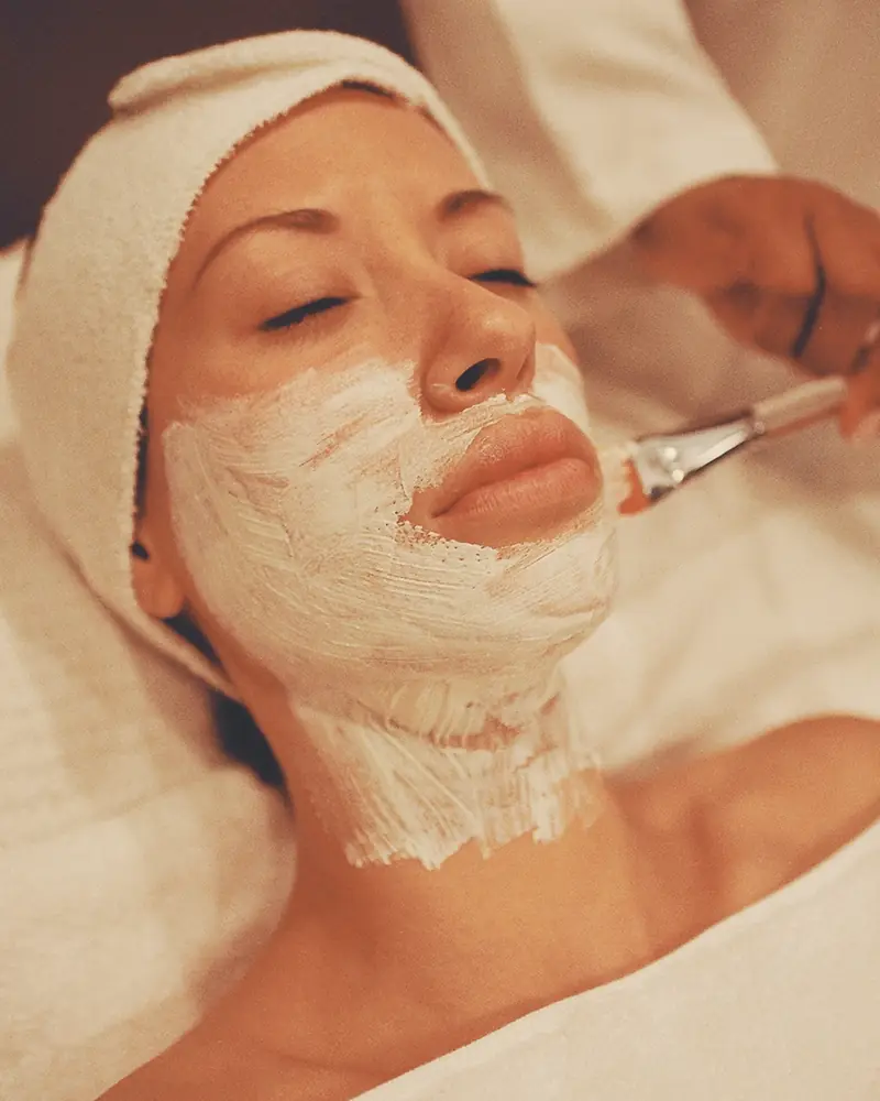 Esteticista aplicando con pincel una mascarilla de arcilla blanca a una clienta